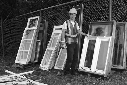 A man wearing a white safety helmet and high-visibility vest is standing outdoors next to a collection of empty white uPVC window frames arranged in a semi-circle. Some of the window frames are propped up against a metal chain-link fence, while others are stacked on the ground. The frames vary in size but are consistent in material and finish, with clear glass panes and a smooth, painted surface. The environment appears to be a yard or storage area, with a gravel or dirt ground and trees or bushes visible in the background behind the fence. The scene is evenly lit, suggesting natural daylight. This image illustrates a component of rubbish removal or garden clearance services, highlighting the process of handling discarded or removed windows and frames commonly encountered in property clearance projects for residential or commercial properties in the Yeading area. The presence of the company name, Garden Clearance Yeading, subtly contextualizes the image within a local outdoor waste removal service. 