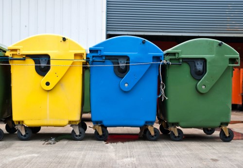 Three large wheeled rubbish bins are lined up on a concrete surface in an outdoor environment. The first bin on the left is bright yellow, with a smooth, matte plastic surface and a black lid mechanism. The middle bin is vibrant blue, made of similar plastic material with a matte finish, featuring a black lid and a secure locking mechanism. The third bin on the right is green, also constructed from sturdy matte plastic, with a matching green lid. All bins are positioned side by side, partially obscuring the lower parts of their wheels, and are set against a backdrop of industrial or commercial surroundings, including a metal-panel wall in white, dark gray, and orange. Natural light illuminates the scene, highlighting the vivid colors and the textured surfaces of each bin. This arrangement reflects typical outdoor waste collection equipment used by services like Garden Clearance Yeading for rubbish management in the local area, emphasizing their capacity for handling garden and general waste disposal.