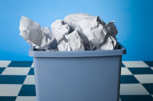 A rectangular, light grey plastic bin filled with crumpled white paper waste, placed on a black and white checkered floor with a solid blue background. The paper waste is piled loosely, with some sheets visibly folded and others more crumpled, occupying most of the bin's interior. The environment suggests an indoor setting, possibly a storage or disposal area associated with garden or general rubbish removal services in Yeading. The overall scene reflects a typical waste collection process, with no other objects or tools visible, emphasizing the bin's contents and the plain surroundings. This image naturally aligns with rubbish removal activities handled by Garden Clearance Yeading, serving as visual confirmation of waste disposal tasks undertaken in outdoor or indoor spaces within the local area.