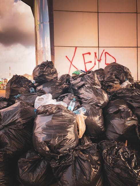 A pile of mixed waste material positioned against a rough, unfinished brick wall and a concrete block wall in an outdoor urban environment. The waste includes cardboard boxes, some flattened and other