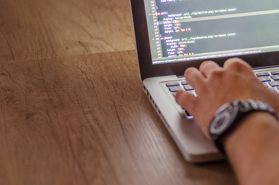 A close-up view of a person's right hand with a silver wristwatch, positioned over a silver laptop keyboard on a wooden surface. The laptop screen displays lines of computer code in a dark-themed edit