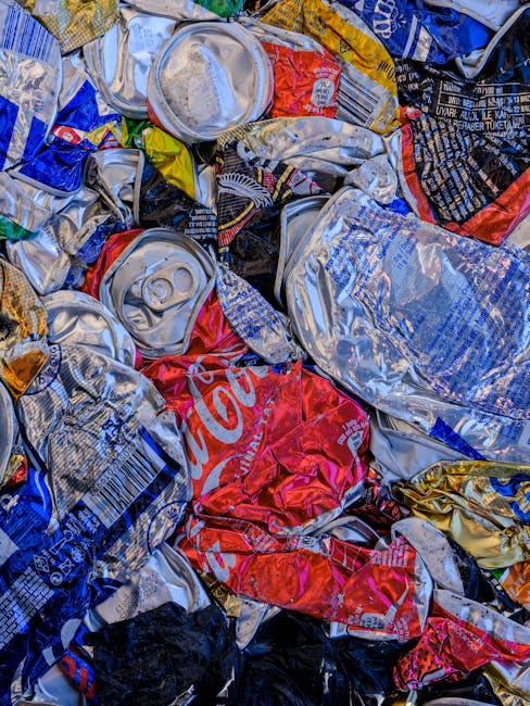 A close-up image showing a tangled heap of crushed and crumpled aluminium beverage cans, predominantly silver with some red, black, yellow, and blue branding visible. The cans display various textures