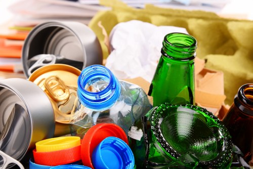 A close-up view of discarded recyclables and waste materials, including crushed aluminium cans with visible pull tabs in various colours such as gold, blue, and yellow, alongside several glass bottles, including a prominent green bottle and a smaller amber bottle, all positioned on a surface with partially visible packaging and paper waste in the background. The scene depicts a mixture of materials typically associated with household rubbish, highlighting items that require proper disposal and waste management in a residential or commercial setting. The image reflects the types of refuse handled by Garden Clearance Yeading during rubbish removal services, emphasizing the need for professional collection and recycling. The environment appears to be an indoor or outdoor area designated for waste sorting or temporary storage before disposal, with natural lighting casting soft shadows and highlighting the textures of metal and glass surfaces.